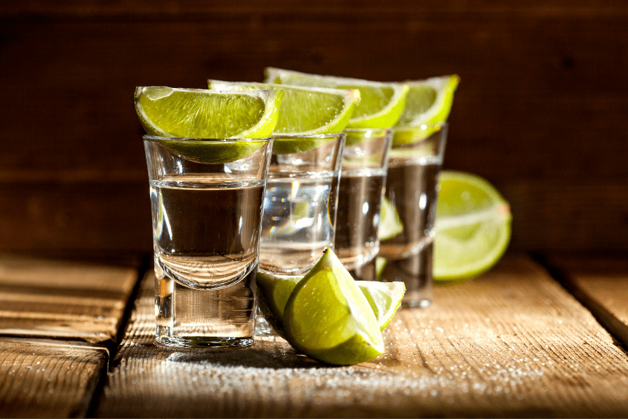 Four small shot glasses are lined up diagonally atop a sunlit table sprinkled with salt. Four lime wedges balance atop each glass