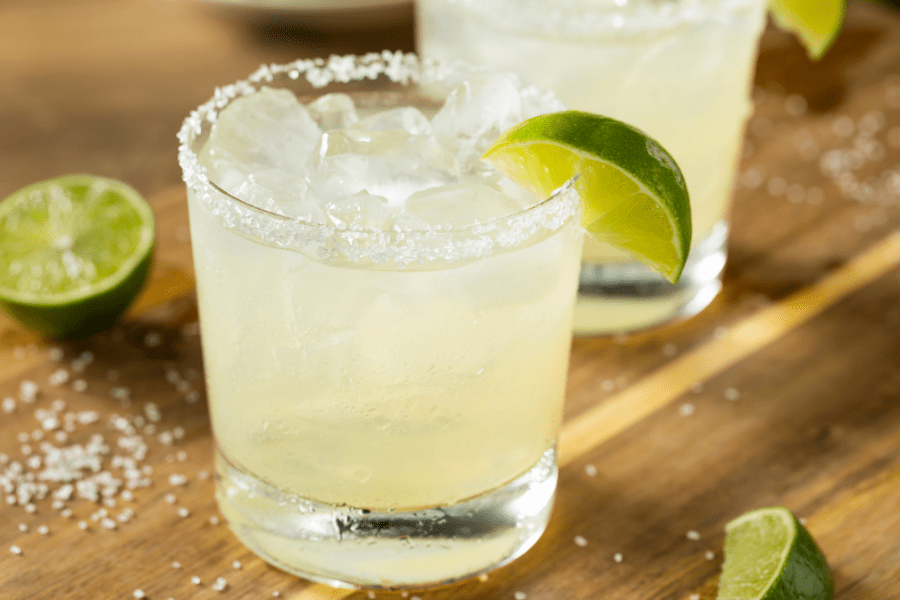 A light wood table top, sprinkled with margarita salt, two rocks glasses filled with a light green margarita, with lime wedges scattered around