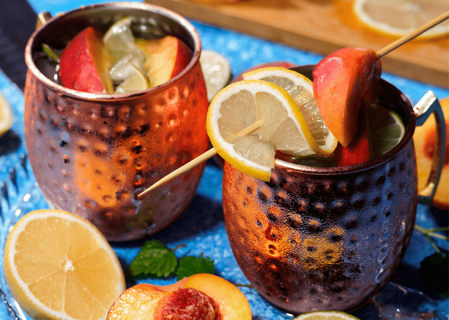 Two copper mugs are seen sitting atop a blue background, filled with a clear liquor and garnished with a skewer of citrus and peach wedges