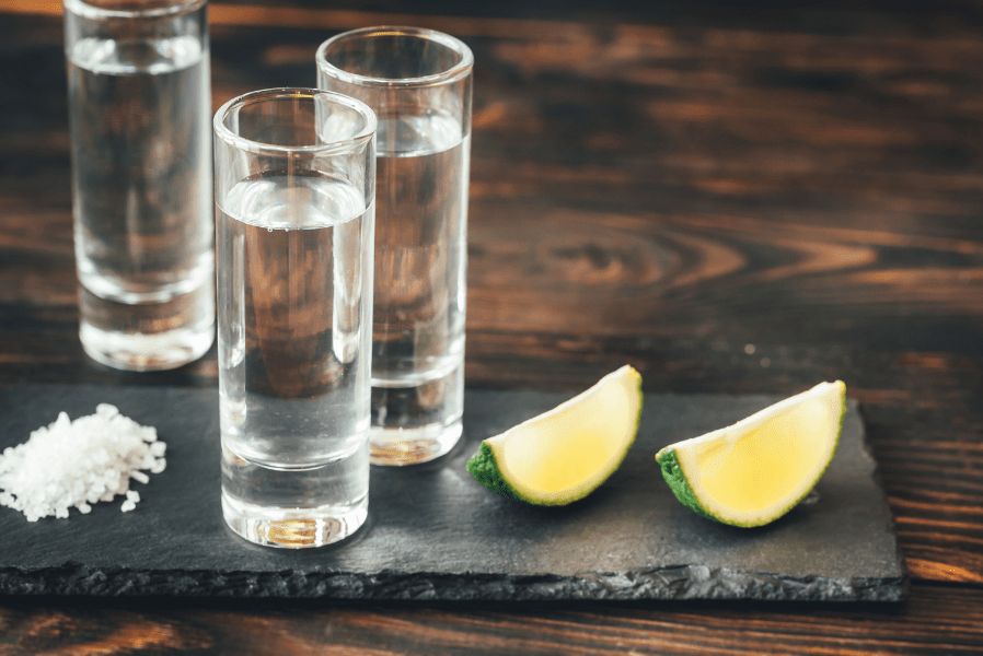 A dark-grain wood table top featuring a pile of large-grain salt, three tall shot glasses filled with clear liquor and two lime slices