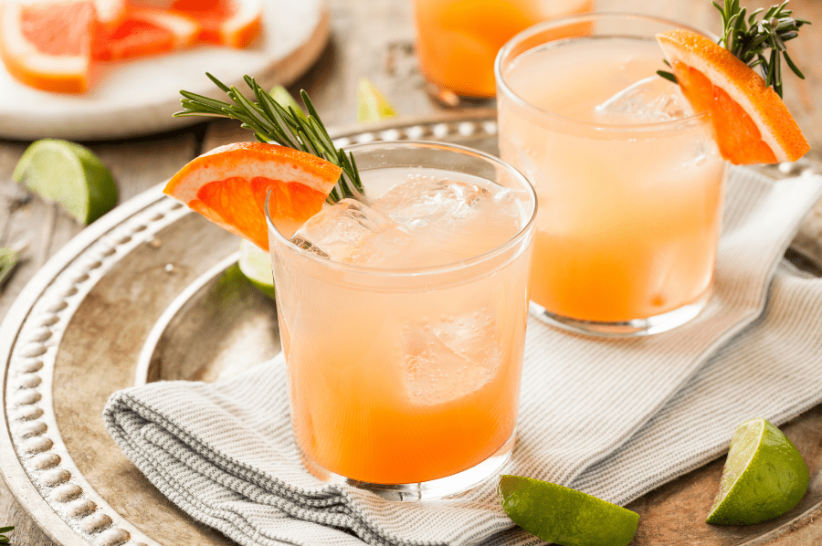 Three pink paloma drinks, garnished with grapefruit wedges and sprigs of rosemary. The glasses are sitting atop a tea towel, silver serving tray, with lime and grapefruit wedges lying about